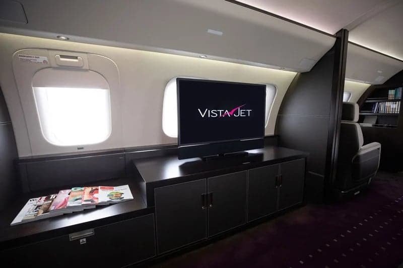 A private jet cabin interior with a large TV screen displaying the VistaJet logo, positioned above a dark wood console.