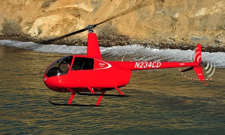 A red Robinson R44 helicopter, registration N234CD, flies over the ocean.