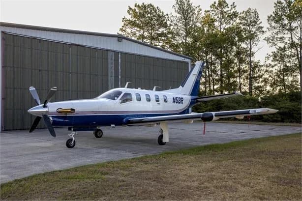 A white and blue single-engine turboprop aircraft, N5BR, is parked on an asphalt ramp next to a hangar with trees in the background.