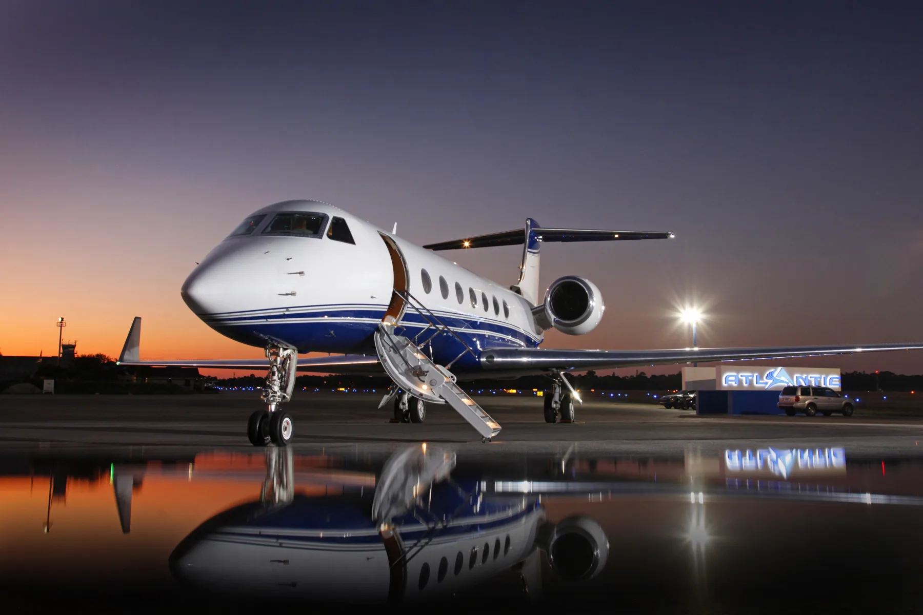 A Gulfstream jet sits on the tarmac at dusk, reflected in the wet surface.