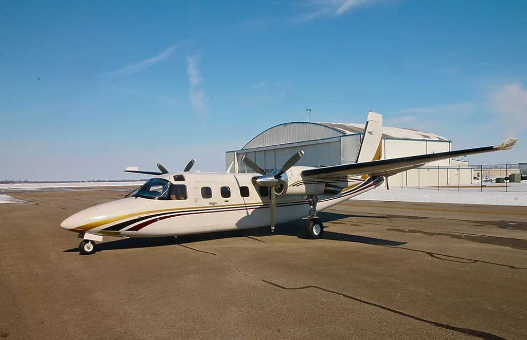 A white twin-engine turboprop aircraft with gold and maroon stripes parked on an airfield with a hangar in the background.