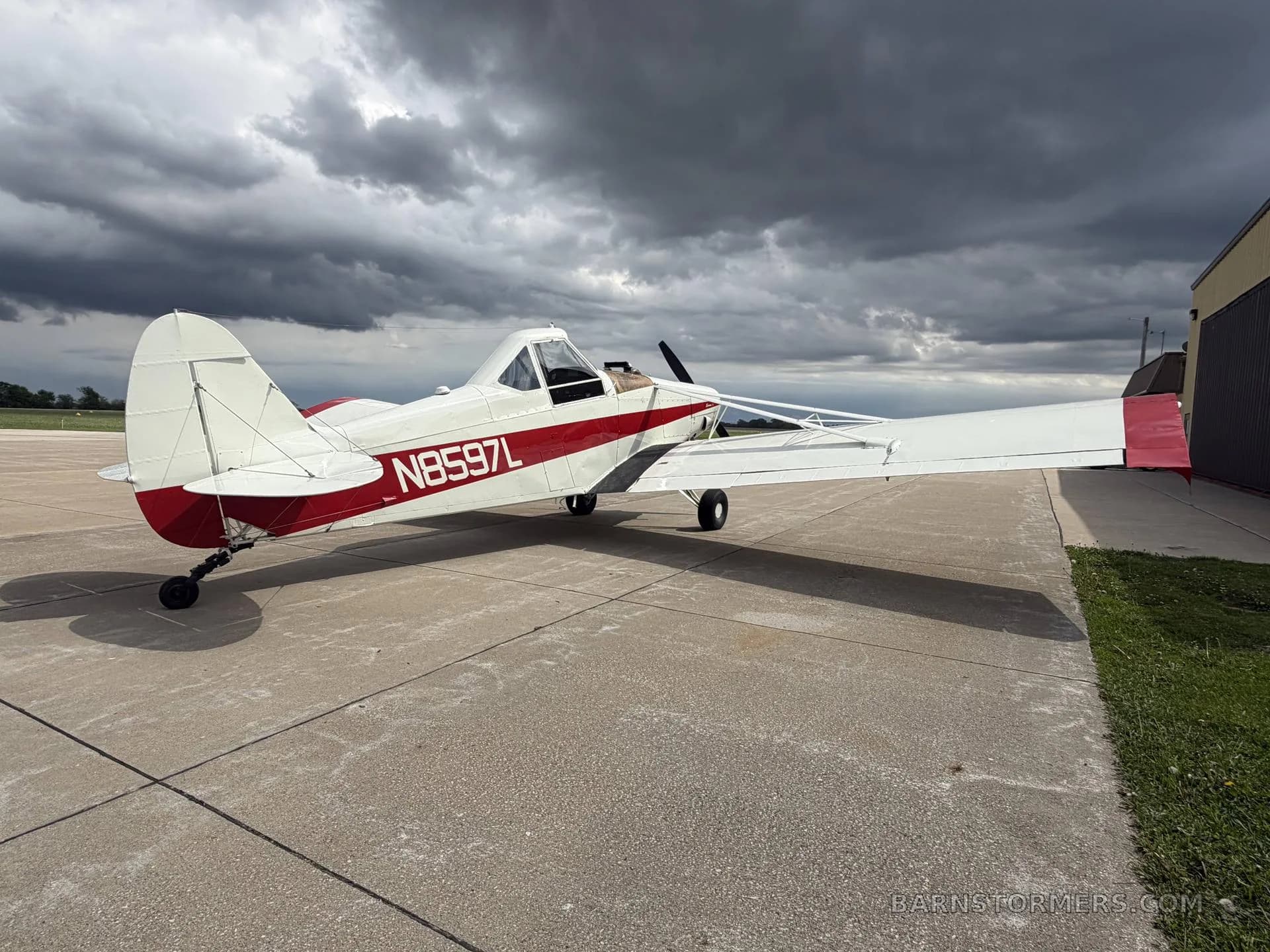 A white and red agricultural aircraft with tail number N8597L, seen from the rear-right side, parked on an airfield under a dark, cloudy sky.