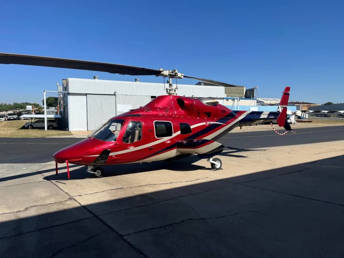A red and white helicopter sits on the tarmac in front of a hangar.
