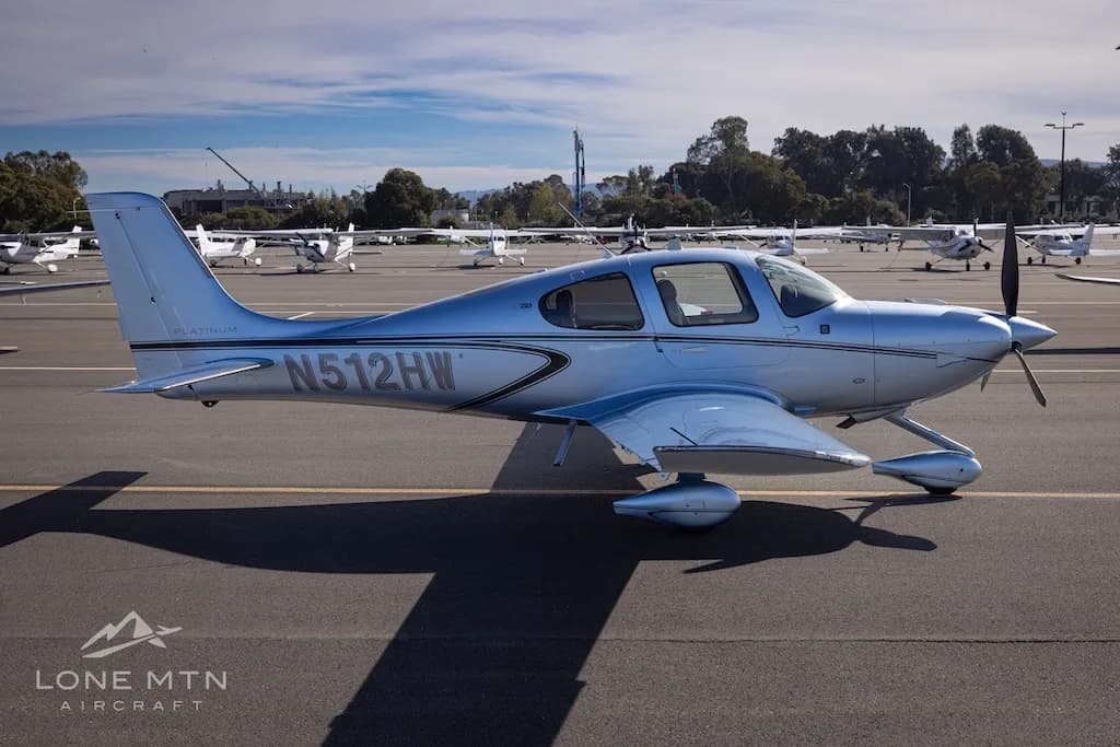 Side view of a silver Cirrus SR22 aircraft, N512HW, parked on an airfield with trees and other aircraft in the background.