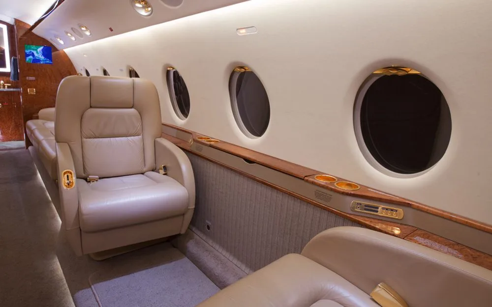 A section of the aircraft cabin with tan leather seats, large oval windows, and wood trim.