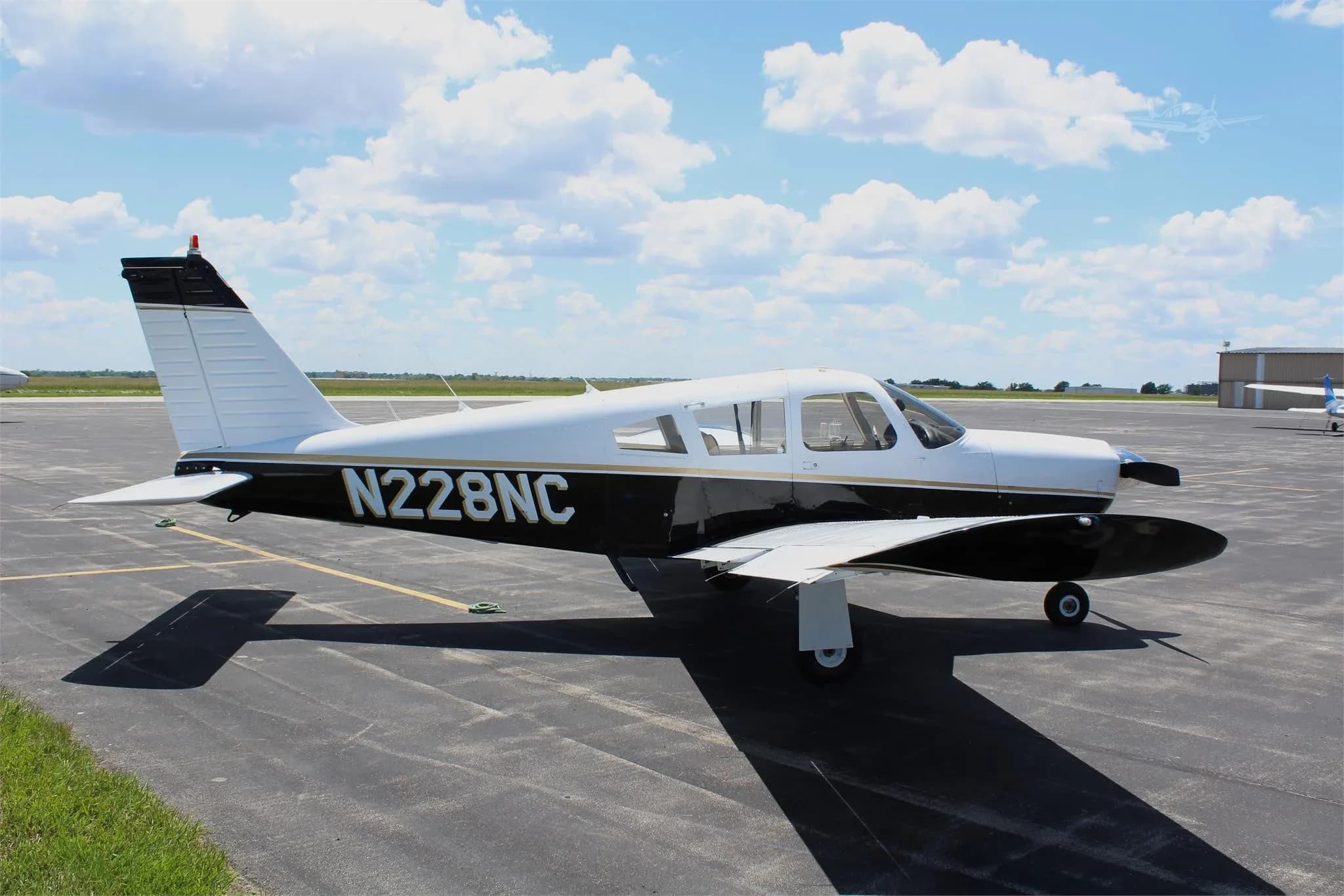 A white and black Piper PA-28 aircraft, registration N228NC, parked on an airport tarmac under a partly cloudy sky.
