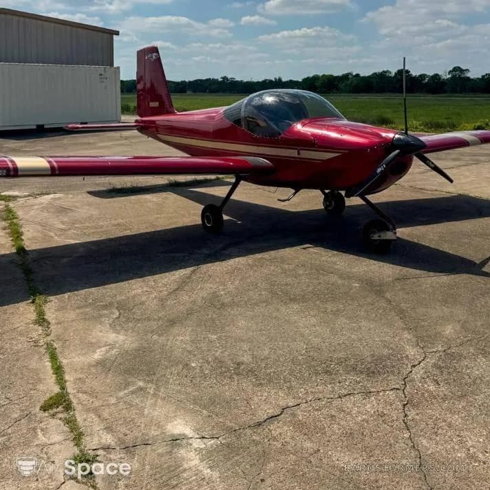 A red Vans RV-6 aircraft with white stripes parked on a concrete tarmac next to a hangar under a partly cloudy sky.