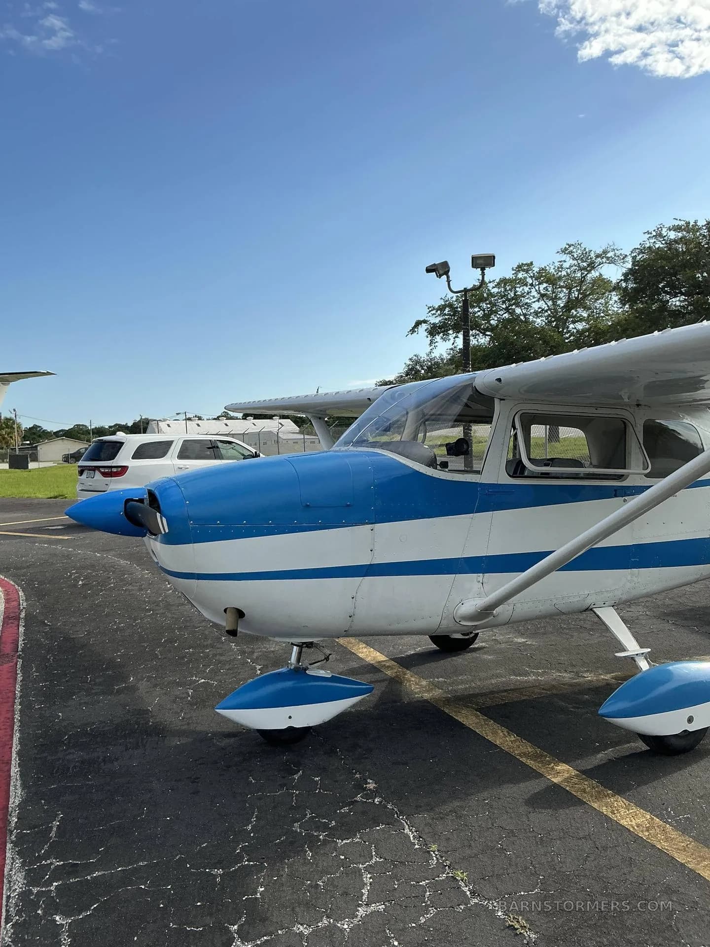 A white and blue Cessna 172 aircraft is parked on an asphalt ramp, viewed from the front-right.
