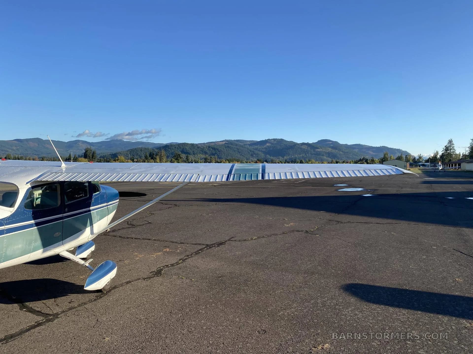 A blue and white Cessna-type aircraft is parked on an asphalt ramp with mountains in the background under a clear blue sky.