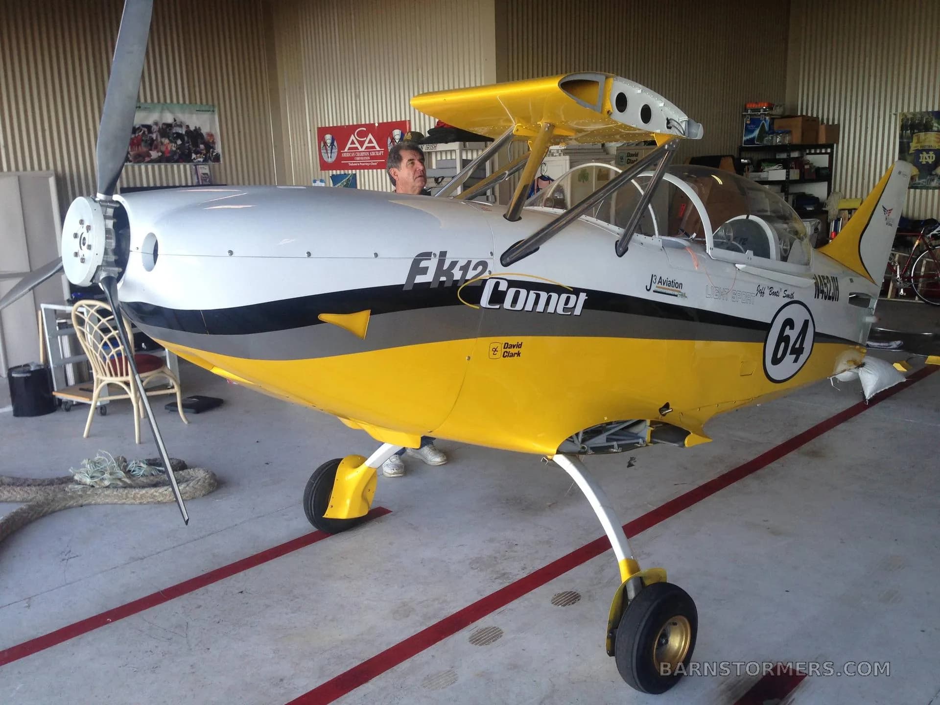 A yellow and white FK12 Comet biplane with '64' on the fuselage, parked in a hangar with a person in the cockpit.