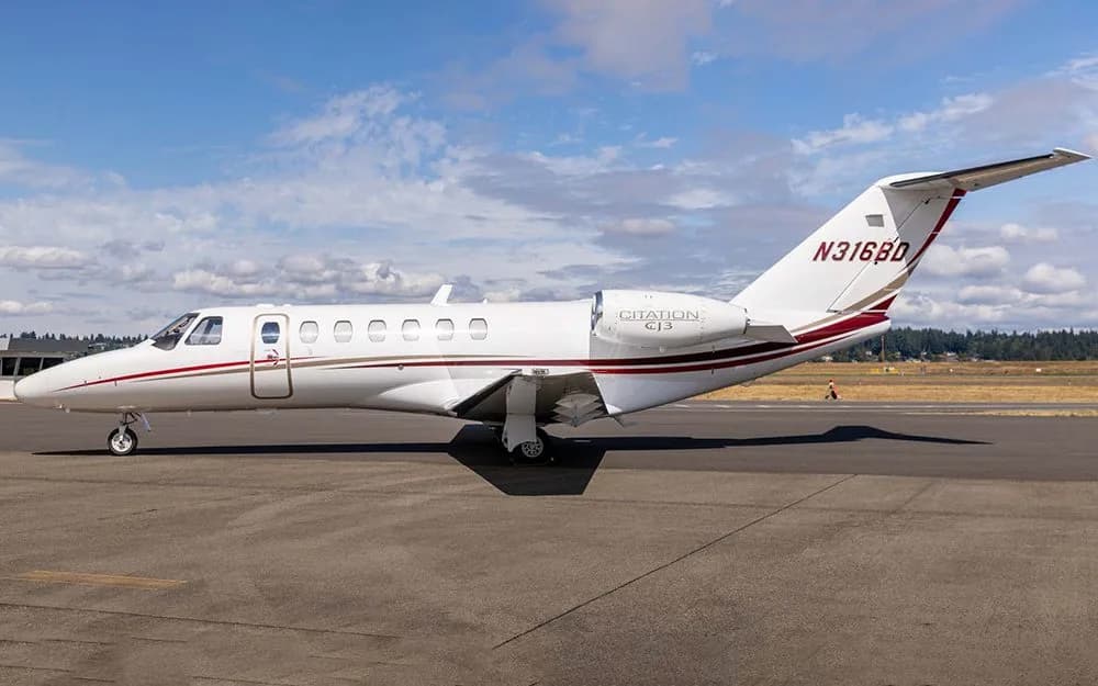 A white Cessna Citation CJ3 business jet with red and gold stripes on the tarmac under a partly cloudy sky.