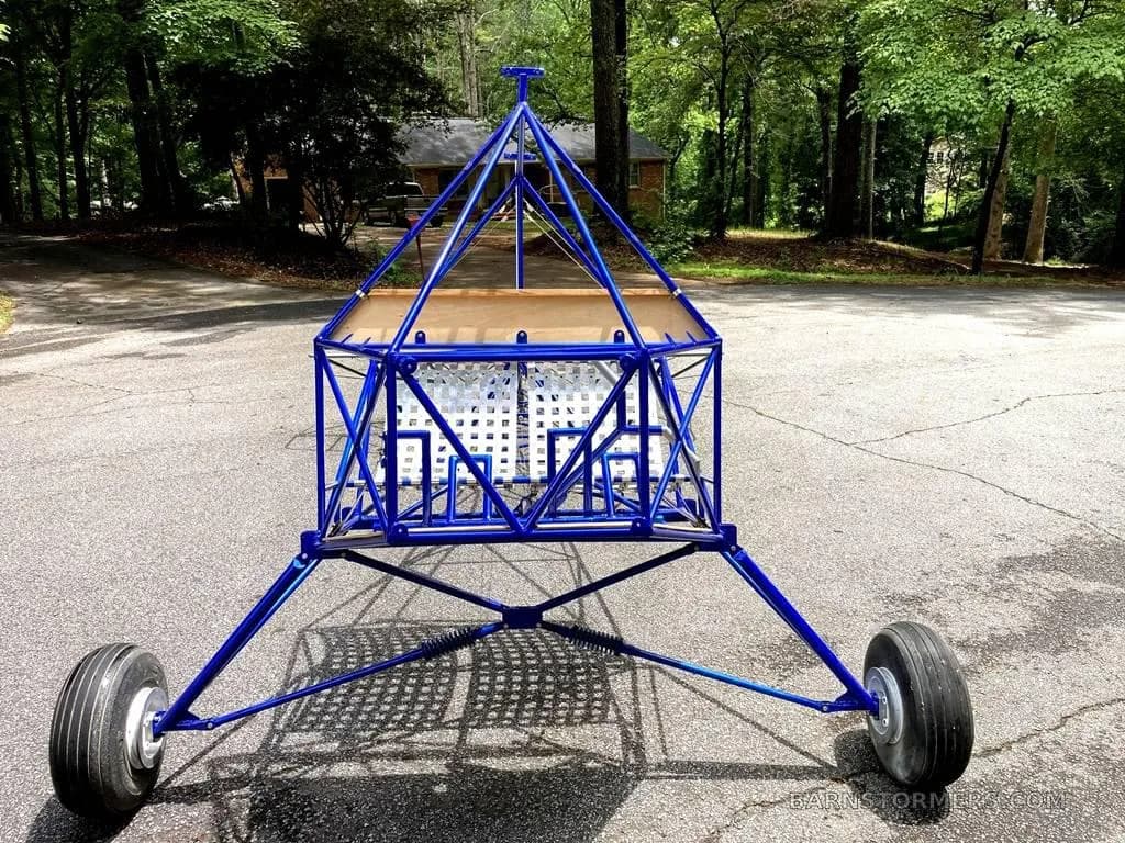 A front view of the blue ultralight aircraft frame, showing the landing gear and a wooden floor.