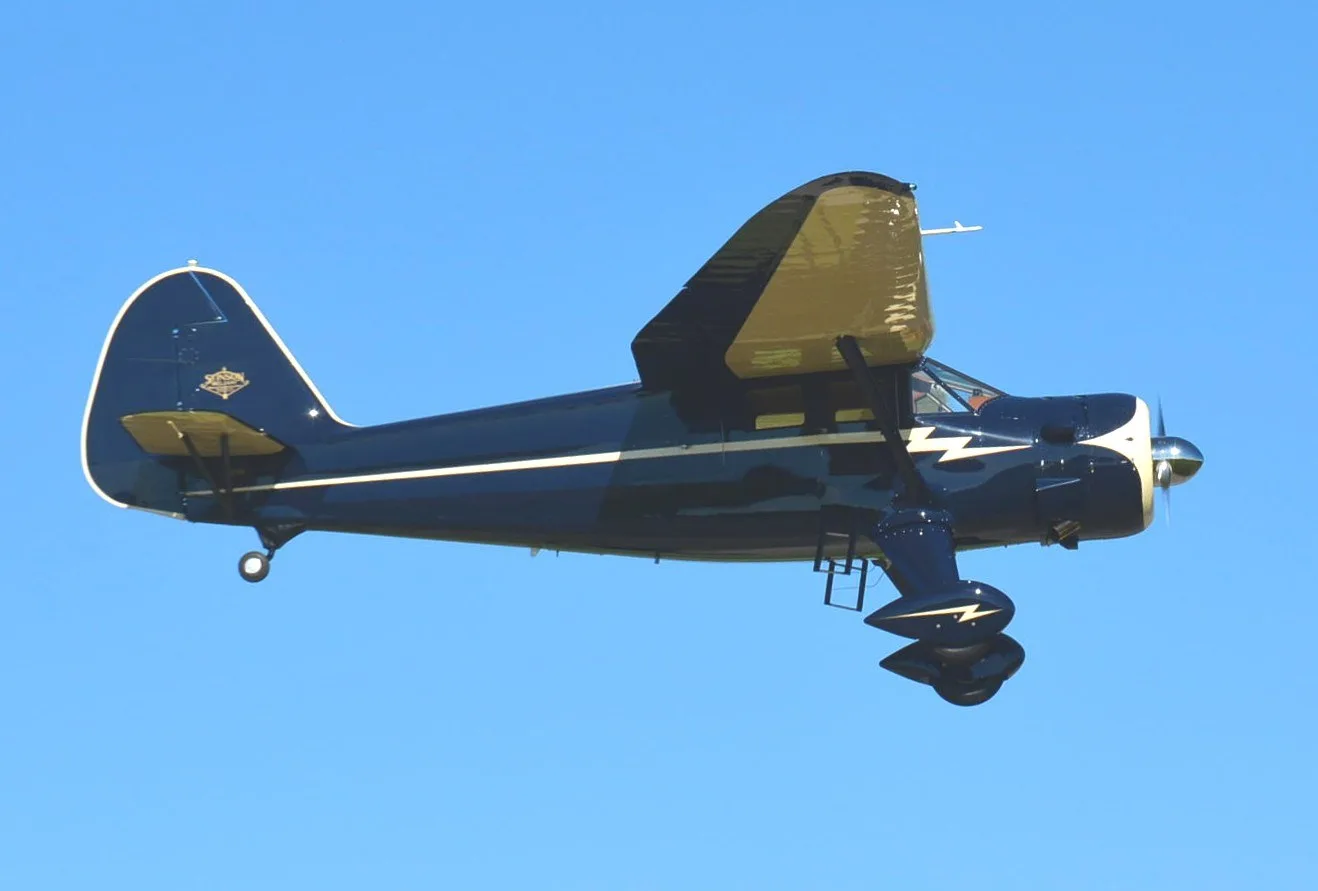 A dark blue and cream vintage aircraft with a lightning bolt design is flying against a clear blue sky.