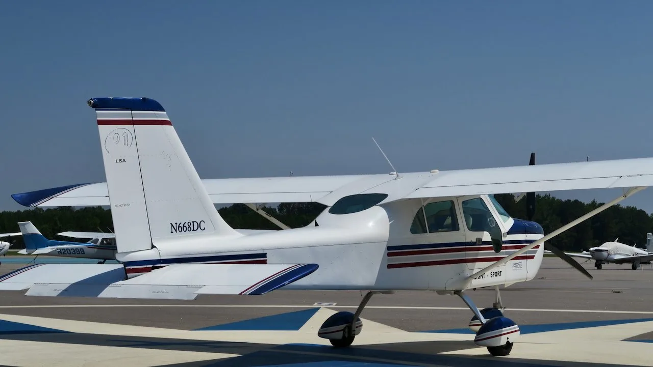 A three-quarter rear view of a white Paradise P1 Light Sport aircraft with blue and red stripes, parked on an airfield with other planes in the background.