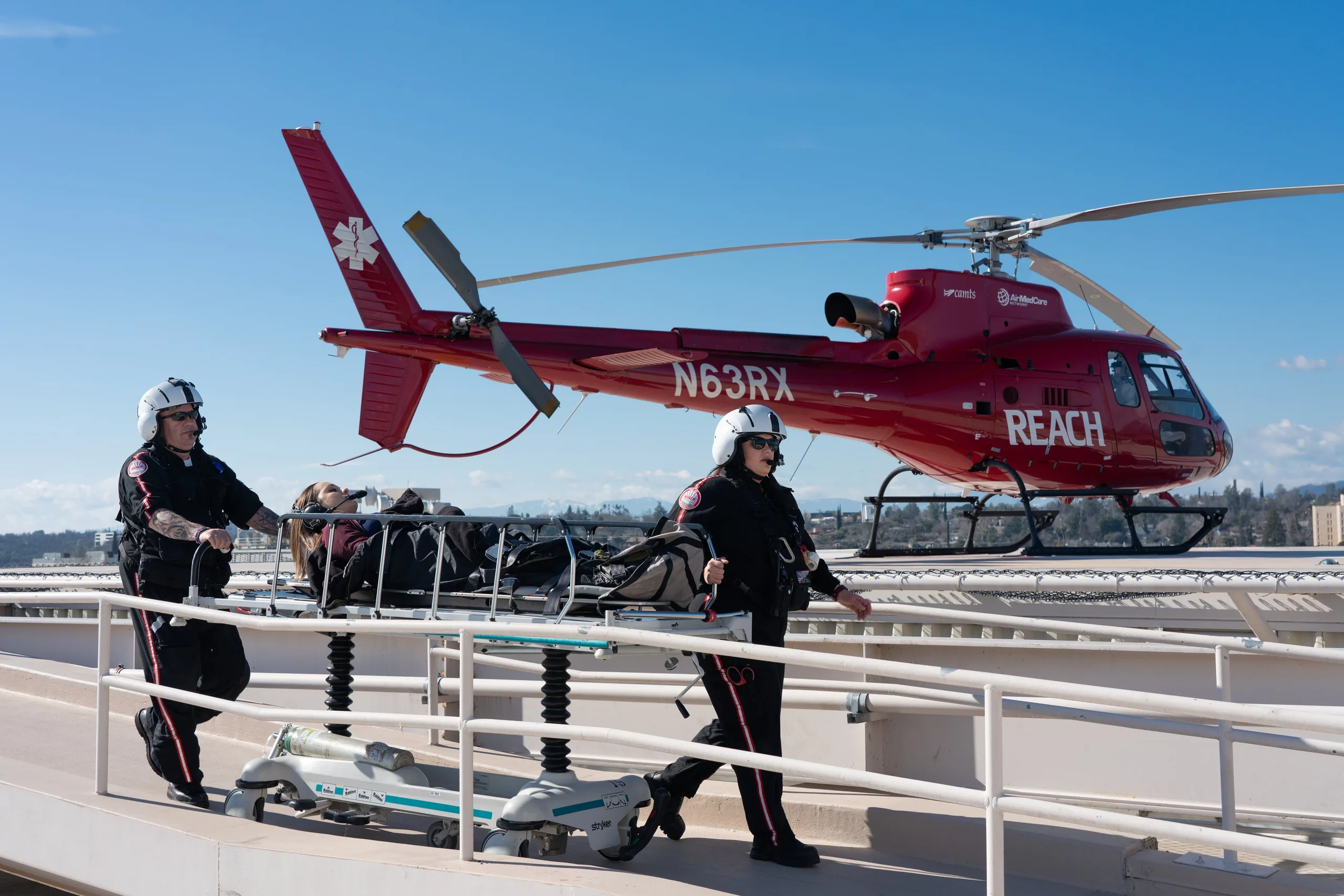 A REACH air ambulance helicopter is parked on a helipad as paramedics transport a patient.
