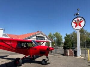 A red Cubcrafters XCub aircraft parked in front of a Texaco sign and building.