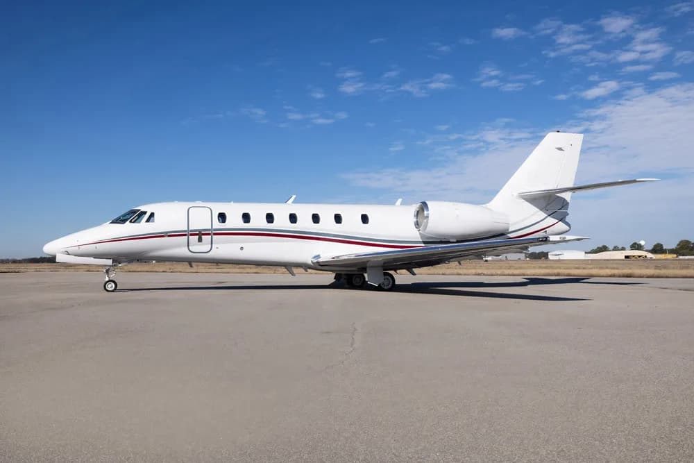 A white business jet with red and grey stripes parked on an airfield under a clear blue sky.