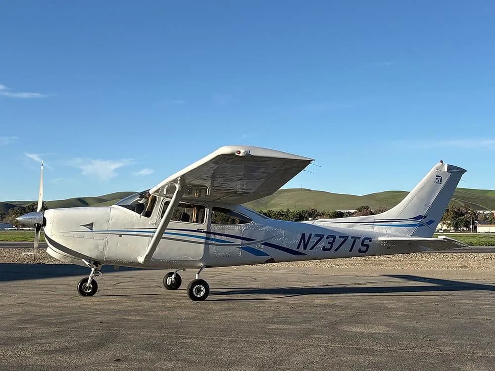 A white Cessna 182 aircraft with blue stripes, registration N737TS, parked on an airfield with rolling hills in the background.