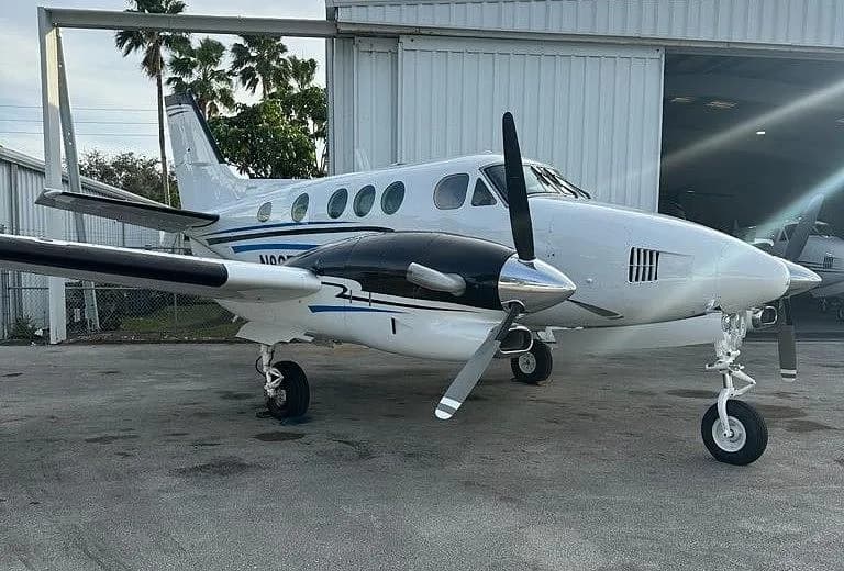 A white and black Beechcraft King Air with blue accent stripes parked on an asphalt apron, partially in front of a hangar.