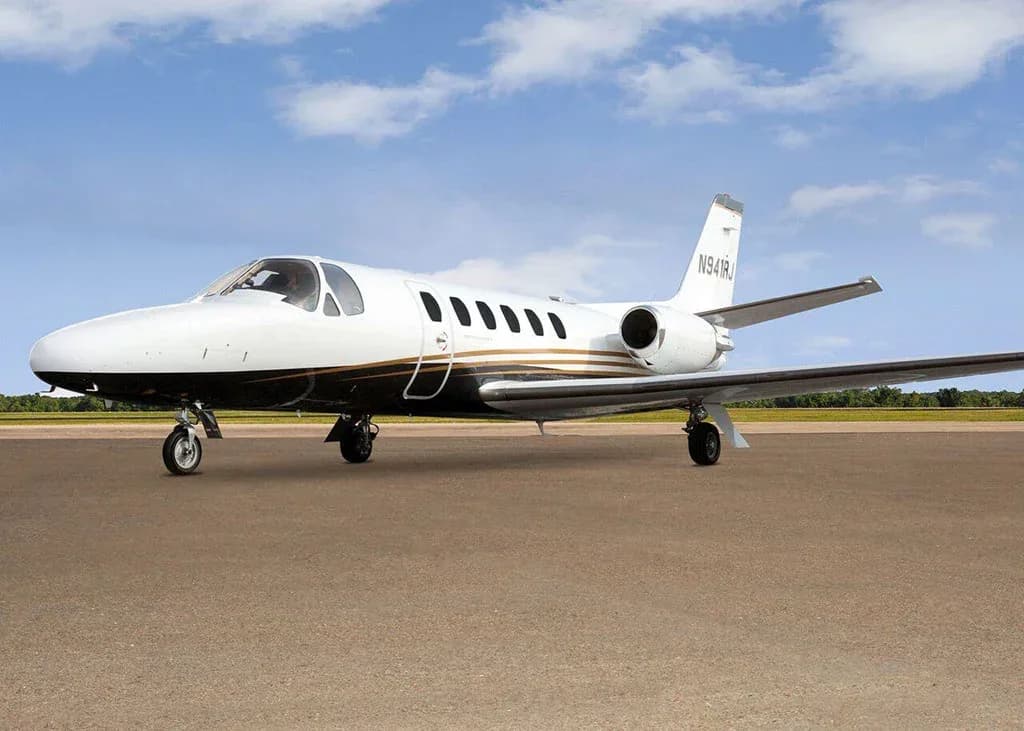 A white and black Cessna Citation business jet N941RJ with gold stripes is parked on a tarmac under a partly cloudy sky, viewed from the front-left.