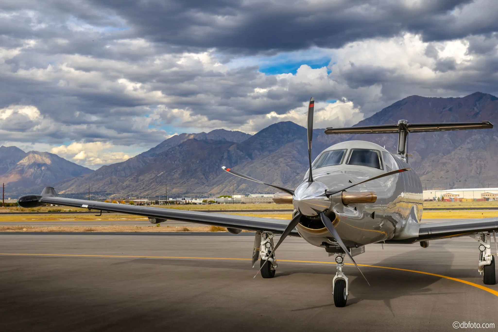 Front view of a silver turboprop aircraft parked on the tarmac with mountains in the background.