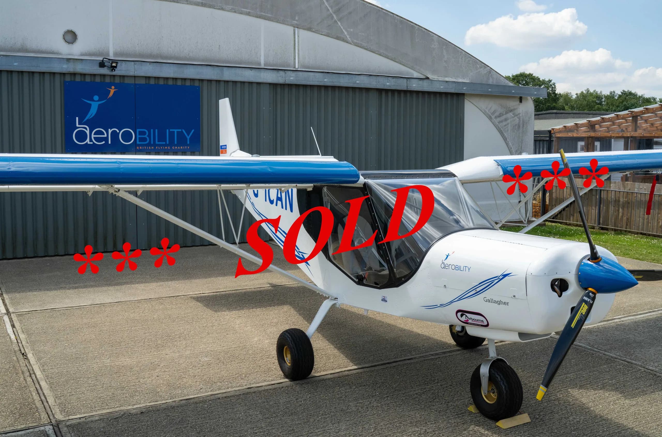 A white and blue light sport aircraft with "SOLD" overlaid, parked in front of a hangar with an "aeroBILITY" logo.