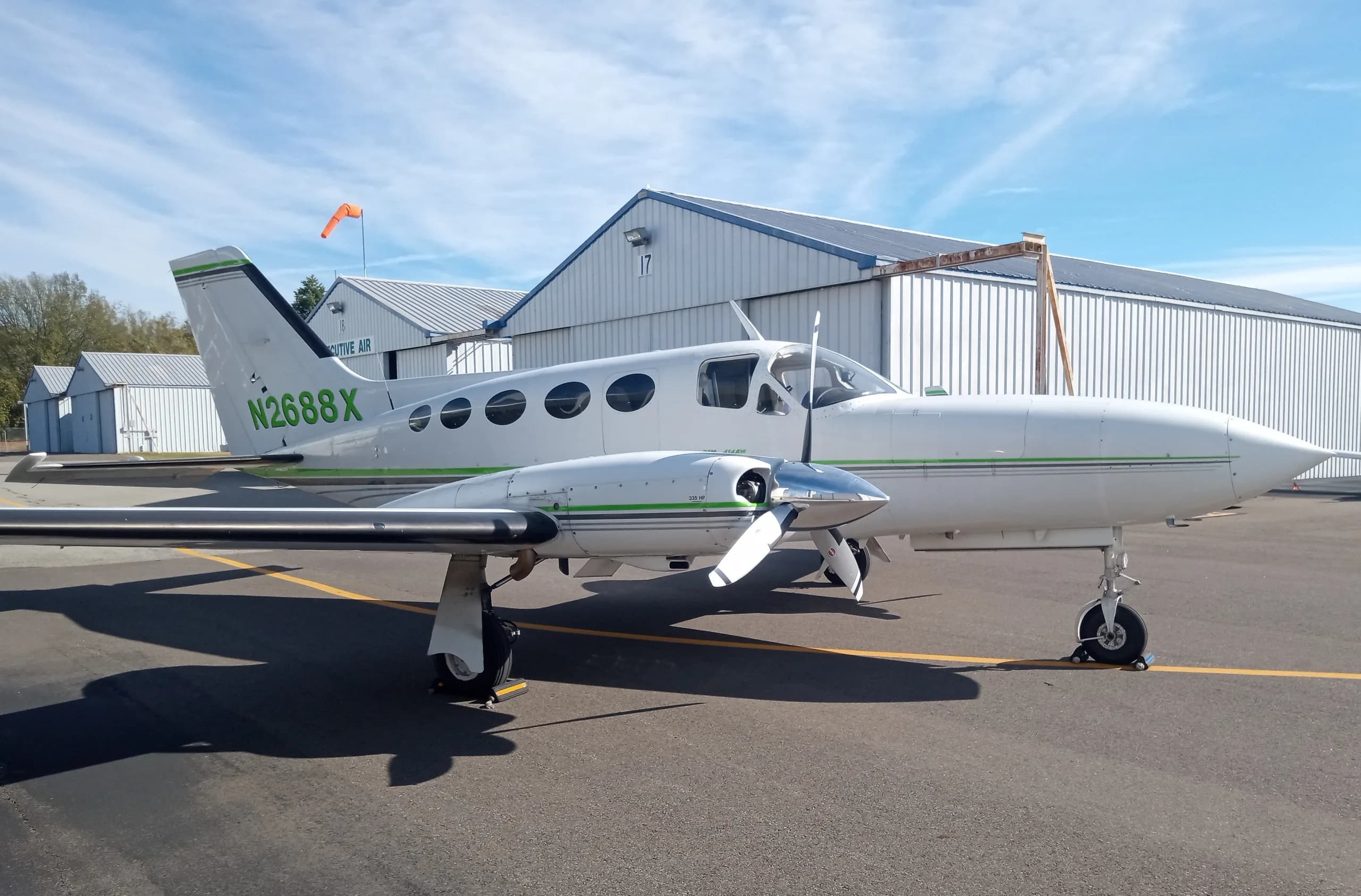 A white twin-engine propeller aircraft with green and black stripes, N2688X on the tail, parked on an asphalt ramp with a hangar and blue sky.