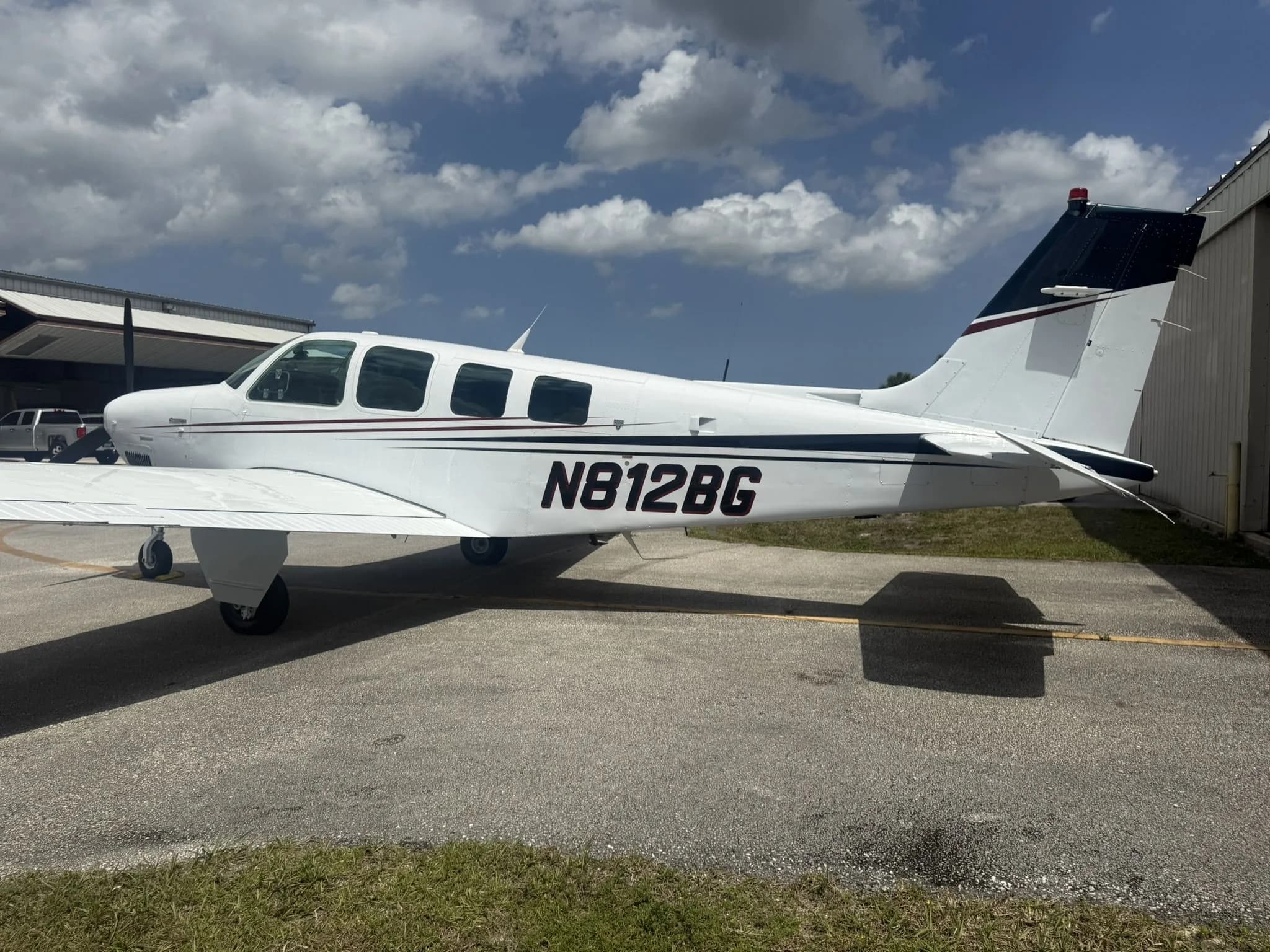 Side profile of a white Beechcraft Bonanza N812BG with dark red and black stripes parked on an airfield.
