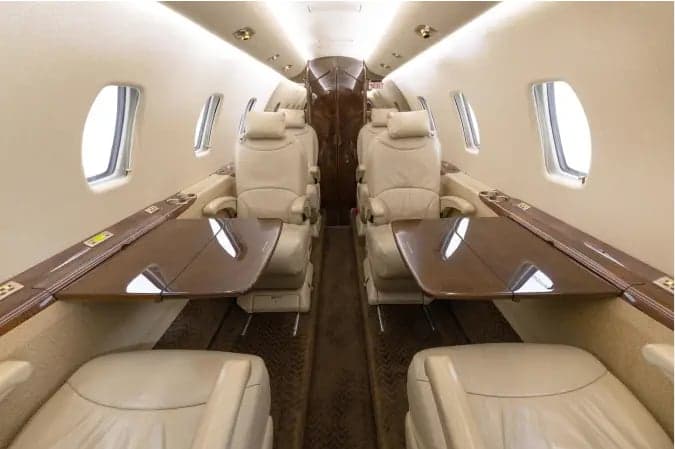 The luxurious cabin interior of a business jet, featuring beige leather seats, fold-out tables, and wood accents.