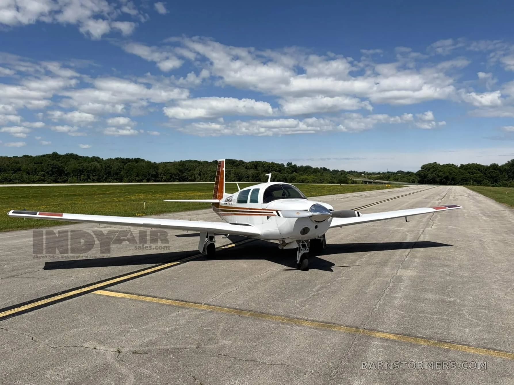A white and brown Mooney M20J aircraft is parked on an asphalt runway under a partly cloudy sky, viewed from the front.