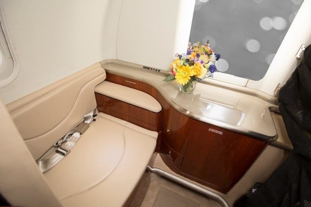 A close-up of a private jet cabin's seating area with a beige seat, wood trim, and a vase of colorful flowers.