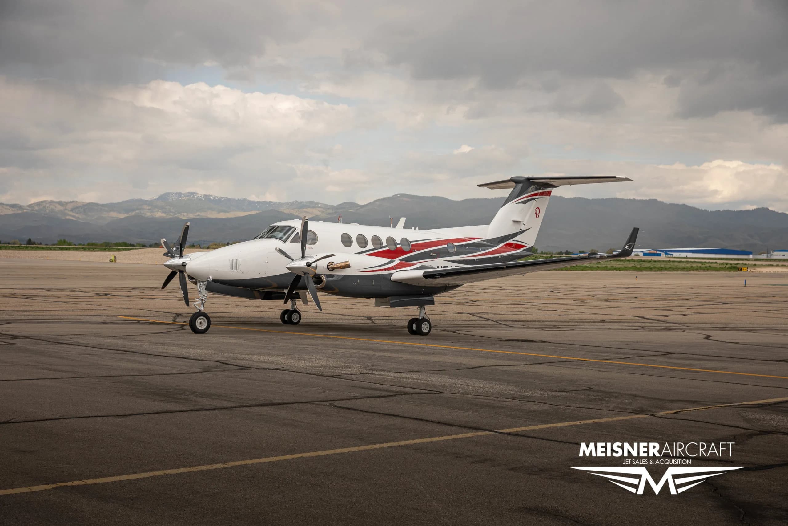 A white, red, and grey twin-turboprop aircraft on an airport tarmac with mountains in the background.