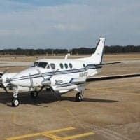 A white Beechcraft King Air twin-turboprop aircraft with blue stripes is parked on an airport tarmac under a partly cloudy sky.
