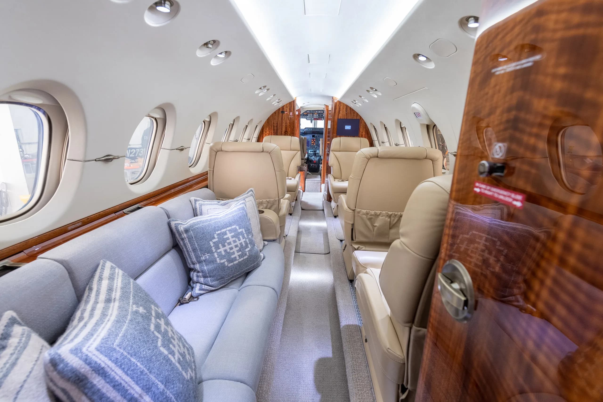 A view down the aircraft cabin aisle, showing a light grey couch with pillows on the left and beige leather seats on the right.