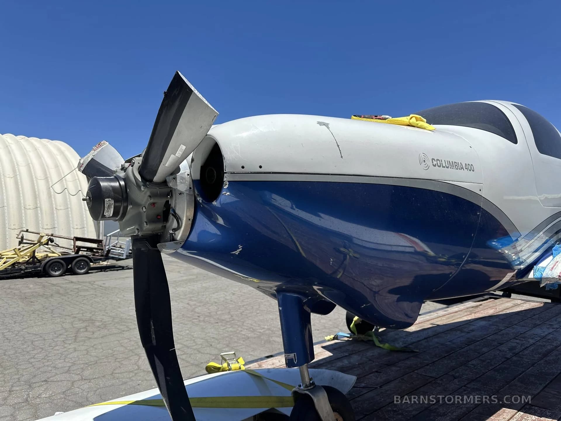 A close-up, low-angle view of the front of a white and blue Columbia 400 aircraft with a damaged propeller, strapped to a flatbed trailer.