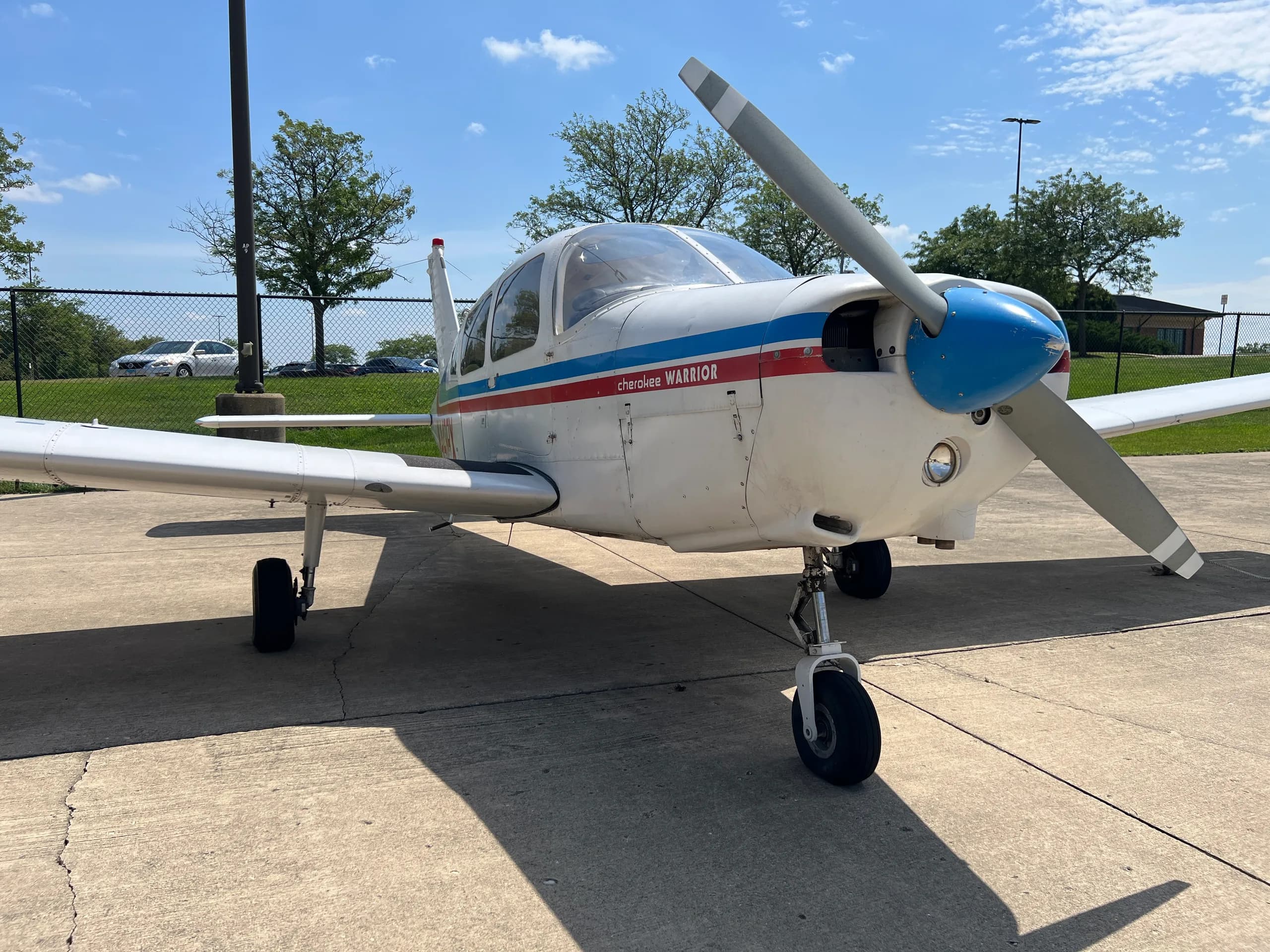 A white Piper Cherokee Warrior aircraft with blue and red stripes is parked on a concrete surface under a clear sky, viewed from the front-right.