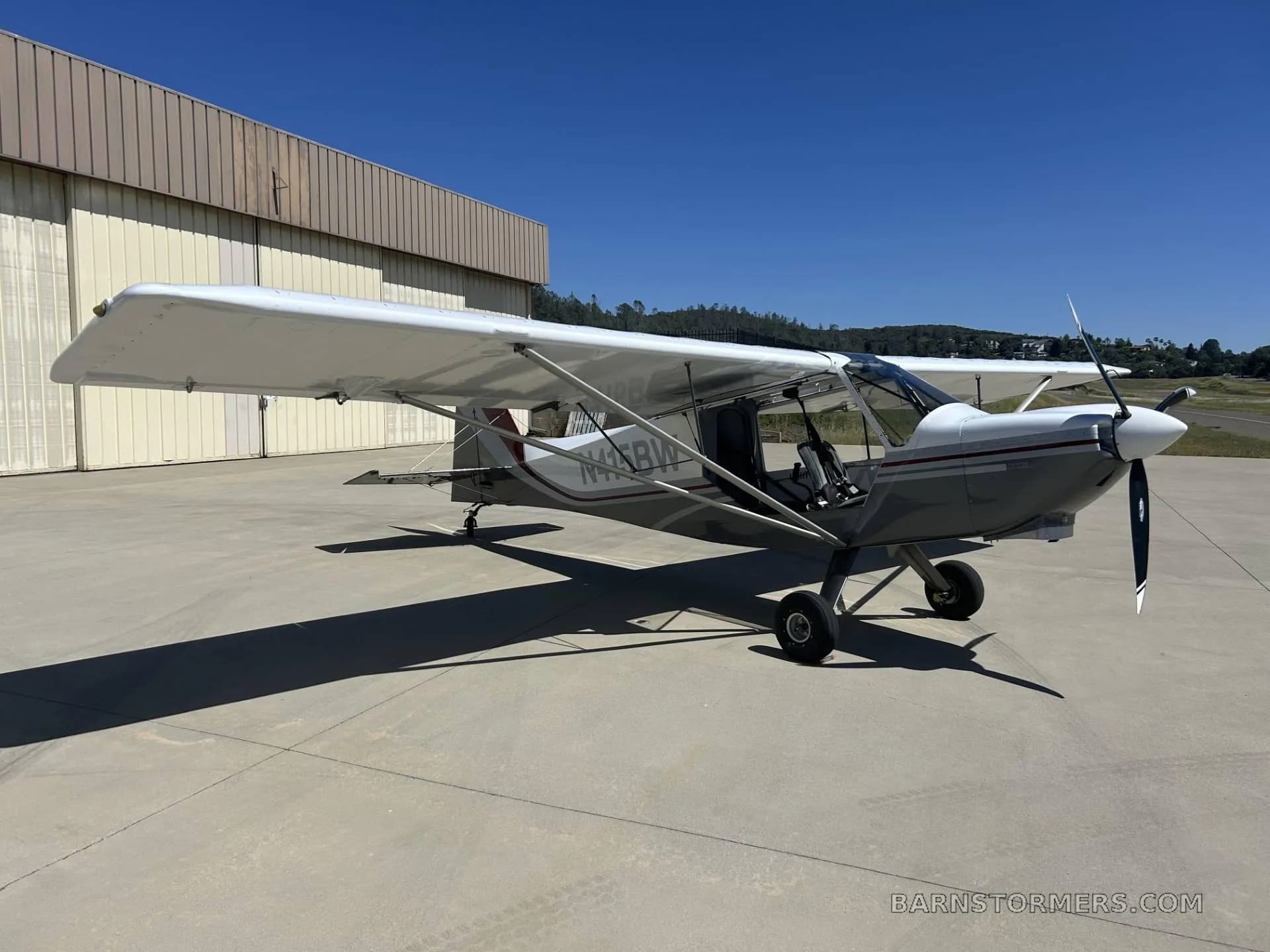 A white and grey Rans S-21 Outbound aircraft, N415BW, parked on a concrete apron next to a hangar under a clear blue sky.