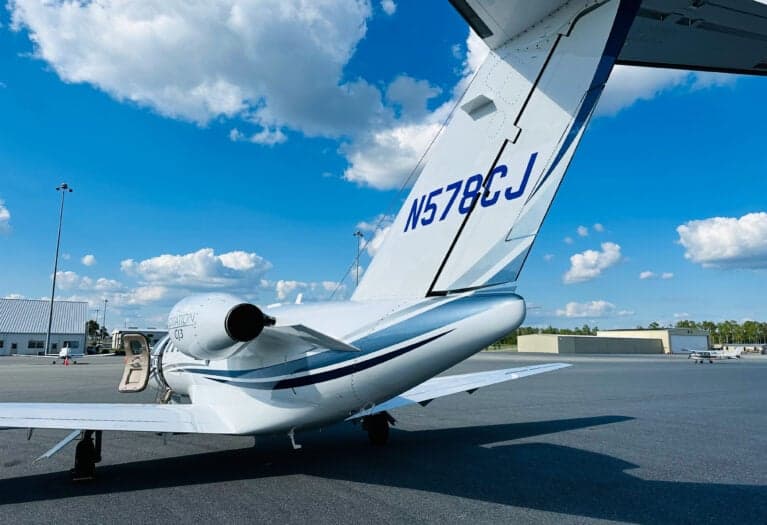 A rear view of a white Cessna Citation CJ3 (N578CJ) with blue and grey stripes, parked on an asphalt tarmac with its main door open.
