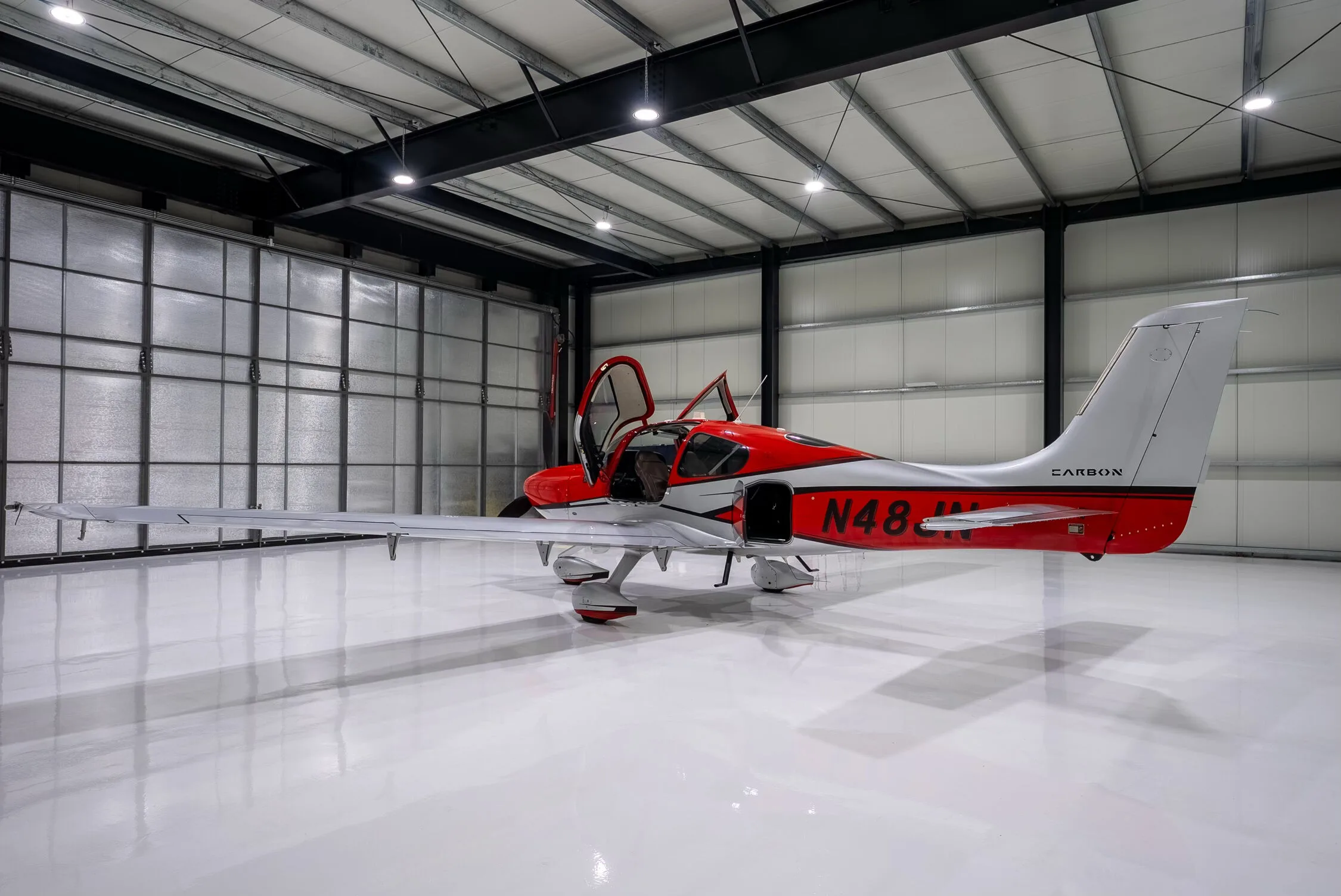 A red and silver Cirrus SR22T Carbon aircraft with registration N48JN and open doors, seen from the rear-left in a hangar.