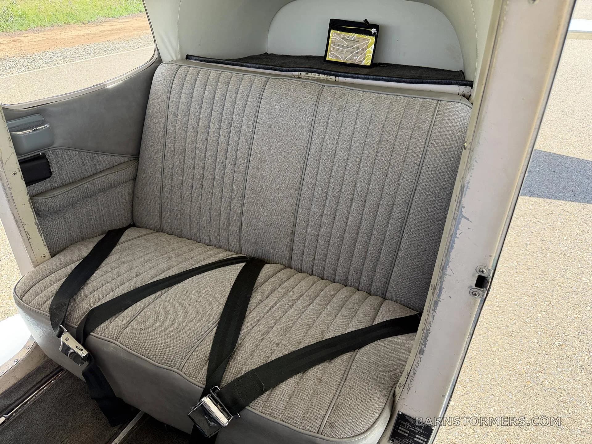 The rear bench seat of an aircraft cabin, upholstered in grey fabric with black seatbelts.
