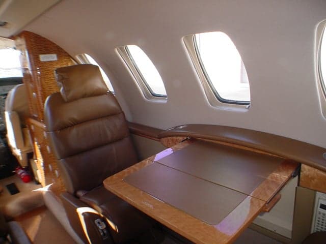 The interior cabin of a private jet, showing a brown leather seat, a fold-out wooden table, and windows.