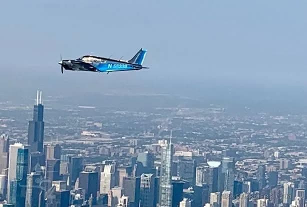 A dark blue single-engine propeller aircraft with registration N 56339 flies over a city skyline on a clear day.
