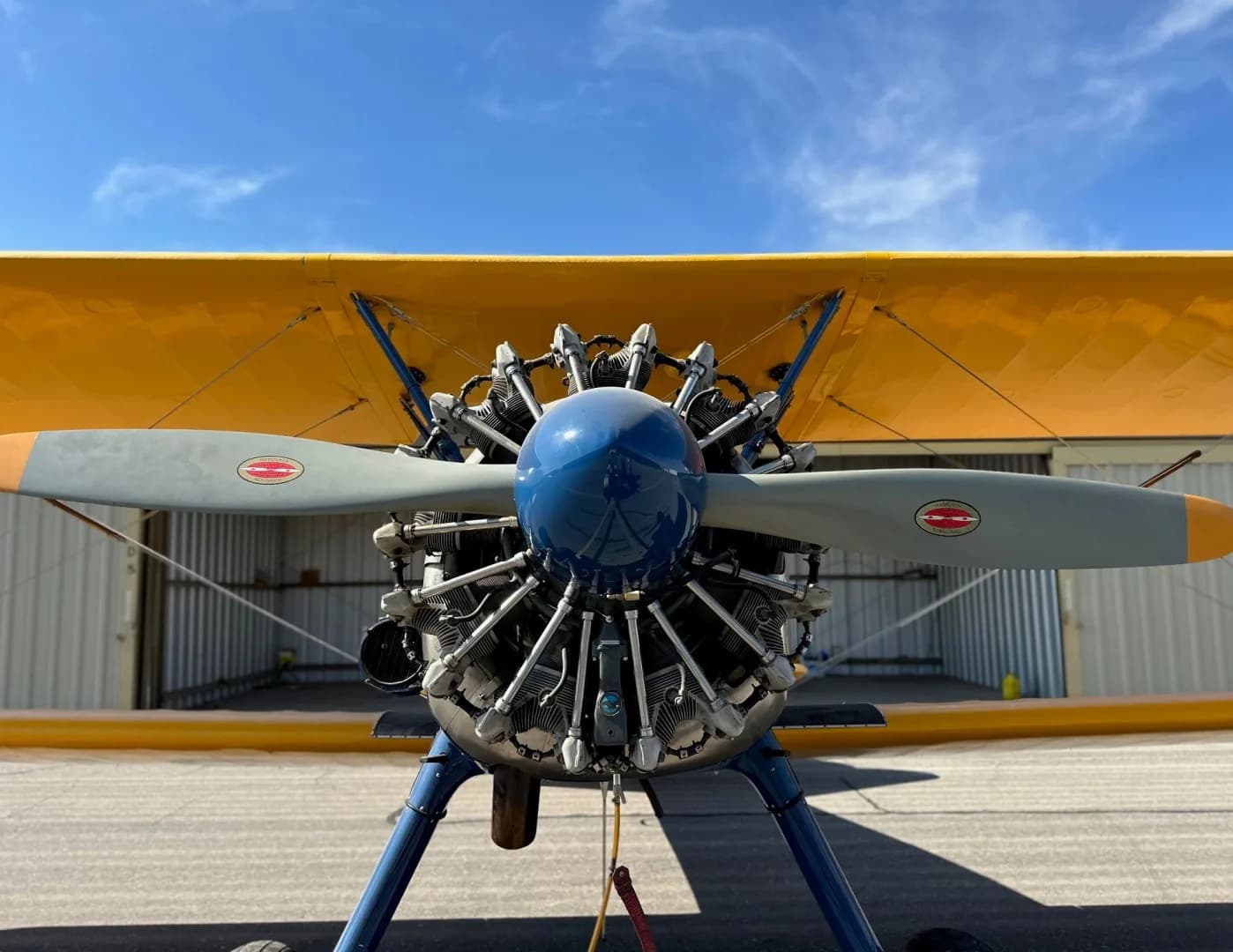 A head-on view of a blue and yellow biplane with a radial engine and a two-bladed propeller, parked in front of a hangar.