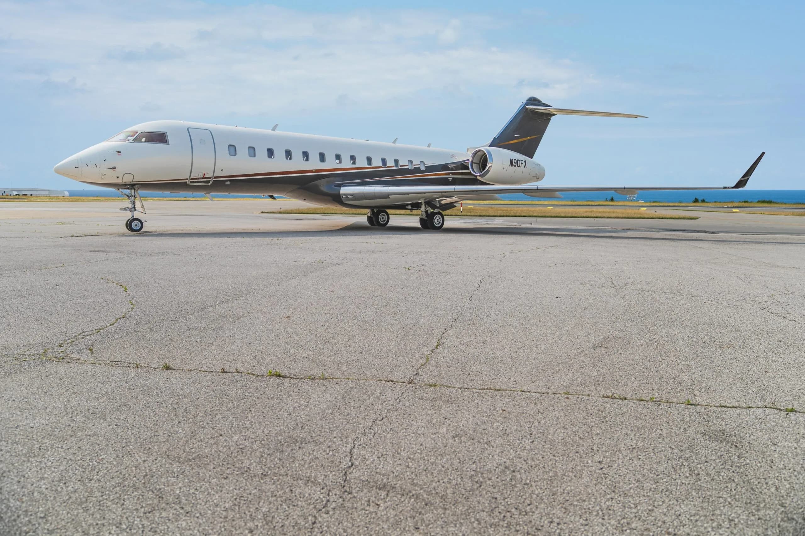 A Bombardier Challenger 605 business jet (N90FX) with a white and dark grey paint scheme parked on an airfield with the ocean in the background.