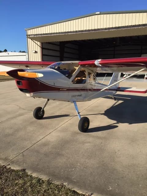 A white and red light sport aircraft is parked on a concrete tarmac in front of an open hangar under a clear blue sky.
