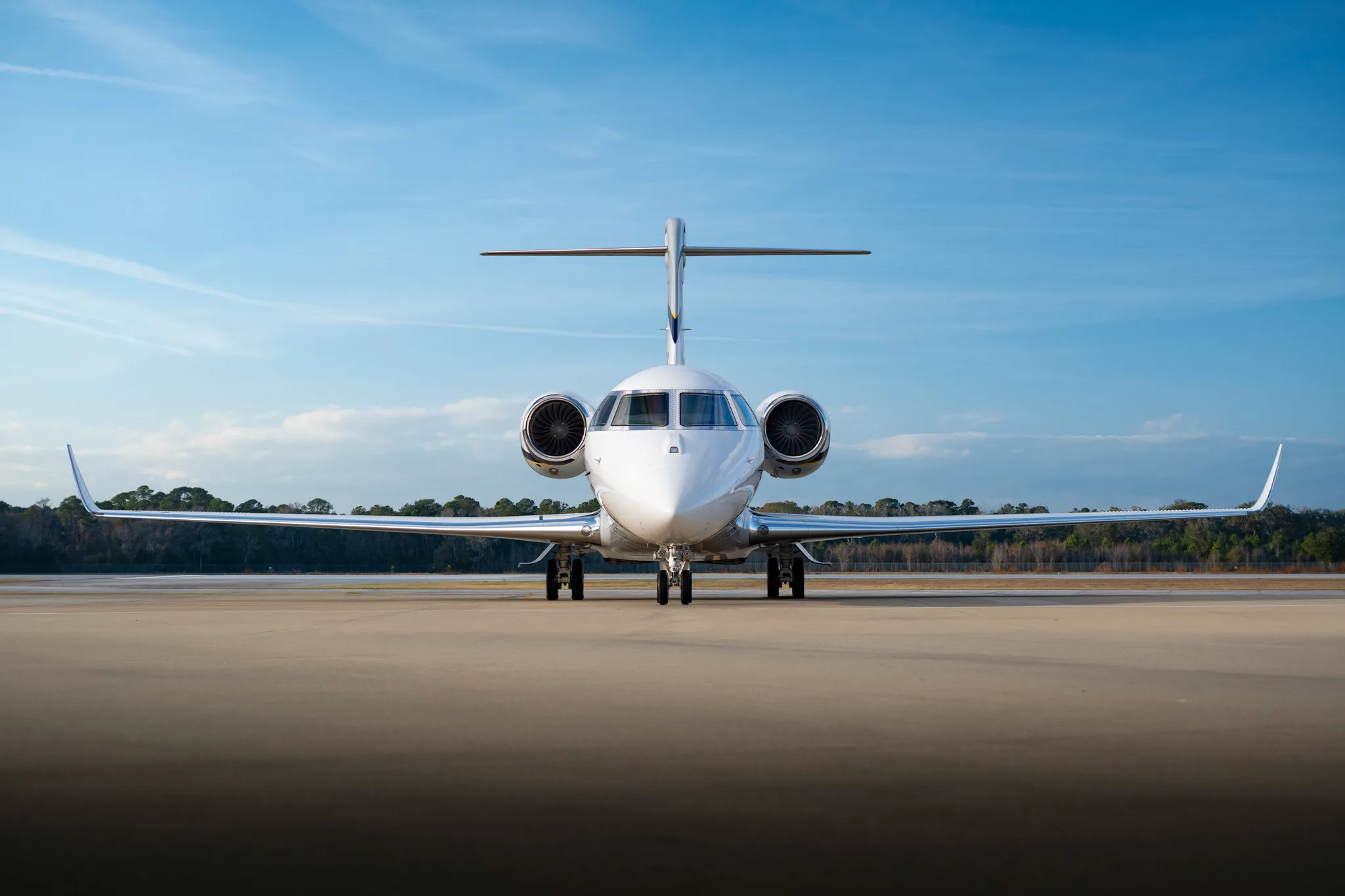 A white business jet with two engines is parked on a tarmac, viewed from the front under a blue sky.