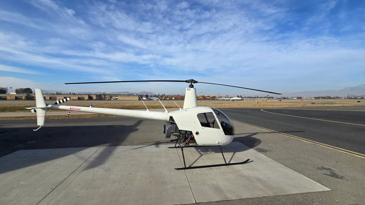 A white Robinson R22 helicopter is parked on a concrete pad at an airport under a partly cloudy sky.