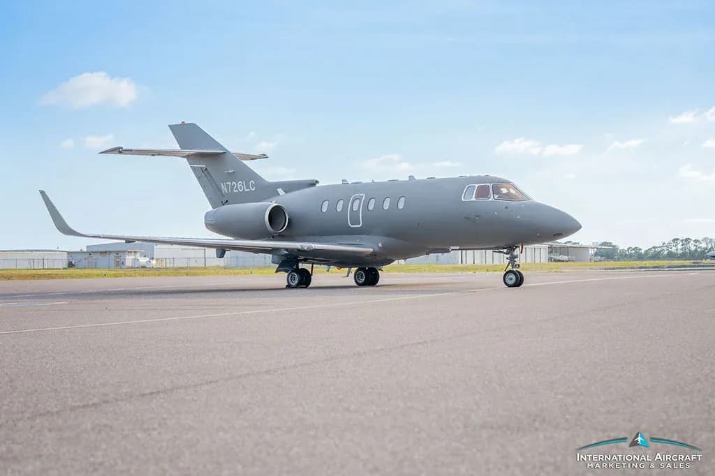 A gray business jet, N726LC, is parked on an asphalt runway with buildings in the background.