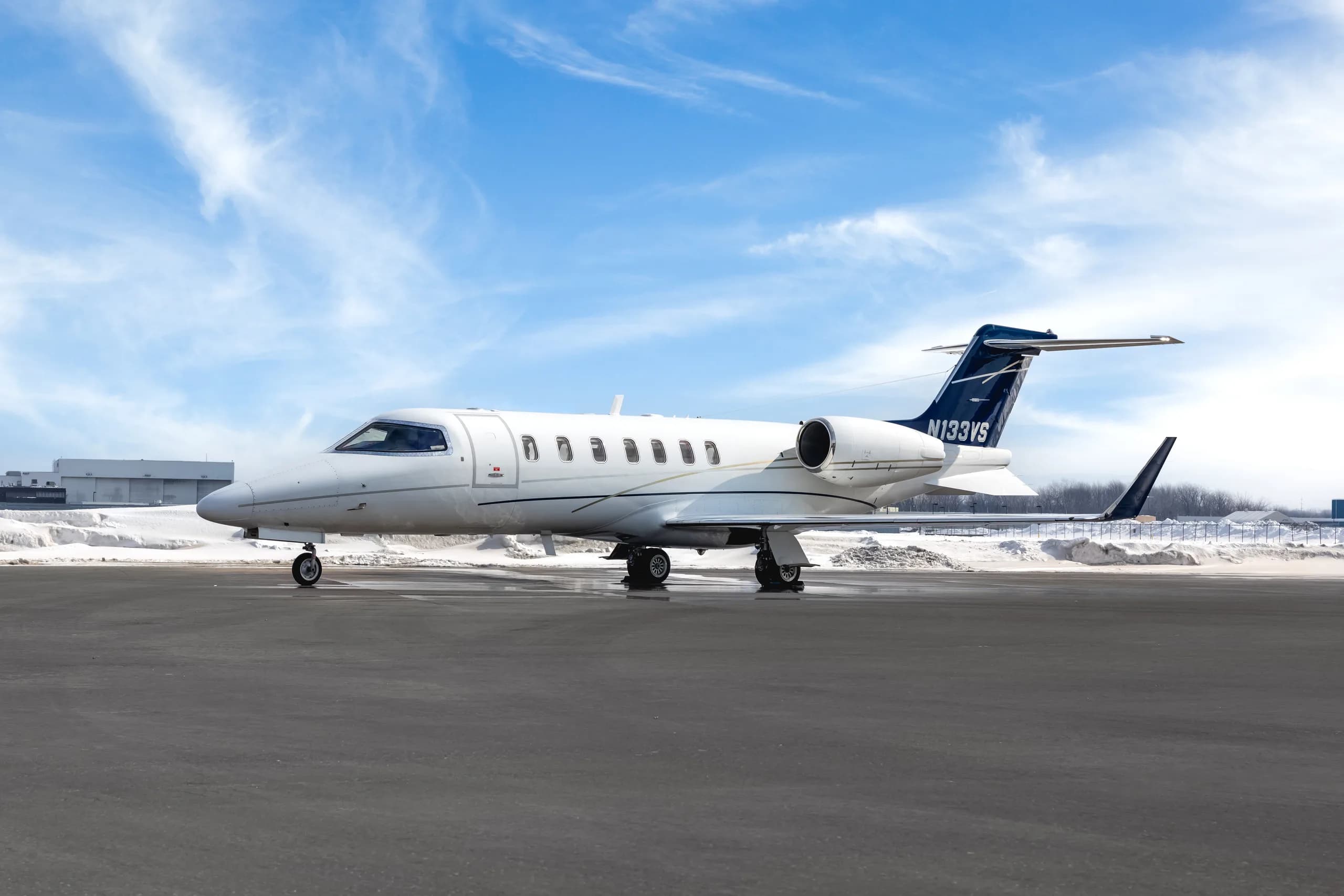 A white Learjet 60 with blue and grey stripes, registration N133VS, parked on a snowy tarmac under a blue sky.