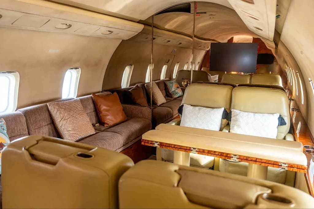 A private jet cabin interior showing a brown sofa, a dining table, and light-colored seats.